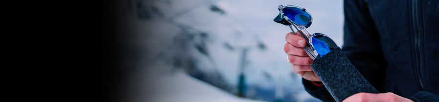 A person puts blue-lensed sunglasses into a dark felt case while standing outdoors in a snowy environment.