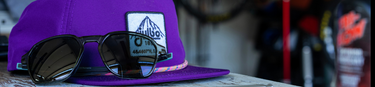 A pair of sunglasses rests on a purple cap with a white mountain patch, placed on a white surface with blurred objects in the background.