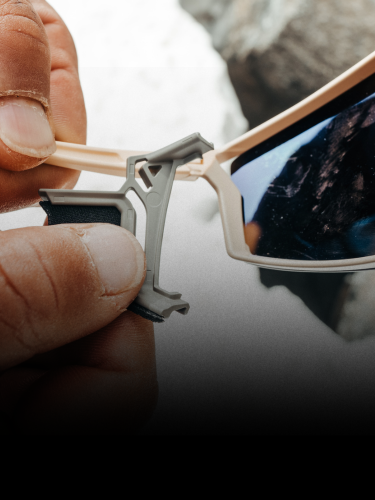 A person attaches or adjusts a metal clip on the lens of beige-framed sunglasses, with a rocky outdoor background reflected in the lens.