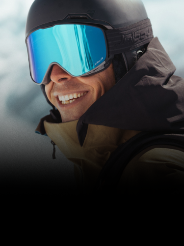 A person wearing a black ski helmet, blue reflective goggles, and a brown jacket smiles outdoors with snow in the background.