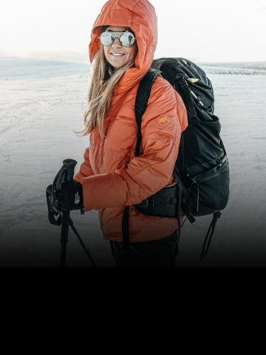 Person wearing an orange hooded jacket, sunglasses, and backpack stands on a snowy landscape holding trekking poles.