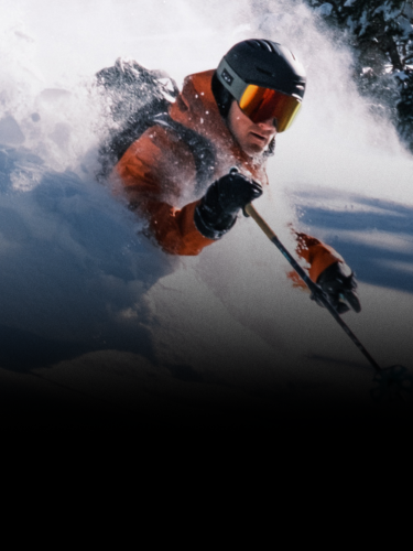 A skier in an orange jacket and helmet moves through deep powder snow, with snow spraying up around them.