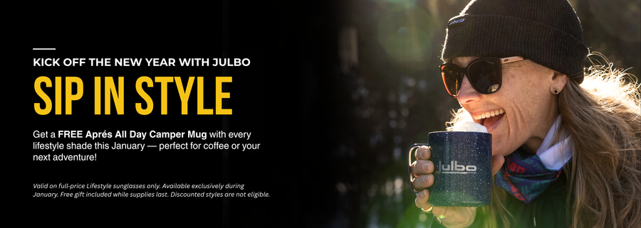A woman wearing sunglasses and a beanie smiles while holding a blue Julbo mug with coffee outdoors in sunlight.