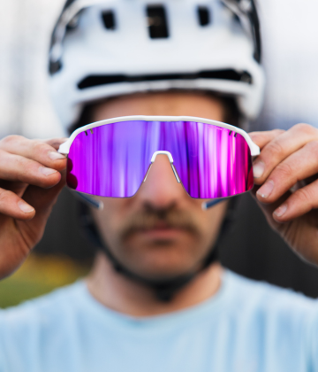 A person wearing a white helmet holds up reflective purple sports sunglasses in front of their face.