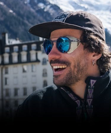 A man wearing sunglasses and a cap smiles outdoors with snow-covered mountains and buildings in the background.
