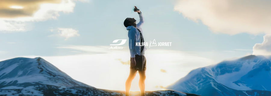 A person stands on a mountain ridge at sunrise or sunset, drinking from a bottle, with snow-capped peaks and clouds in the background. Logos for Julbo and Kilian Jornet appear in the center.