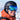 A person wearing a black helmet, blue ski goggles, and an orange jacket is smiling outdoors with snowy mountains reflected in the goggles.