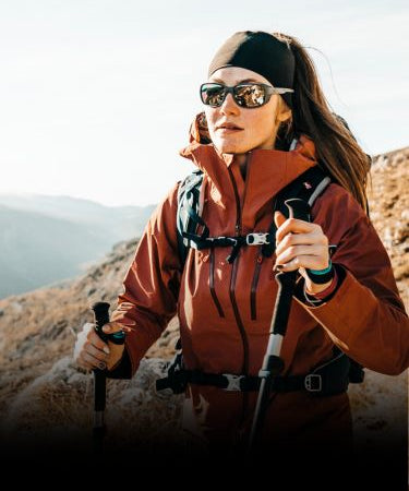 A woman wearing sunglasses and a red jacket is hiking on a rocky trail, holding trekking poles, with mountains in the background.