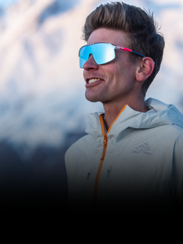 A person wearing reflective sunglasses and a light jacket stands outdoors with snow-covered mountains in the background.