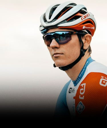 A cyclist wearing a white helmet, sunglasses, and a red, blue, and white cycling jersey looks to the side against a blurred background.