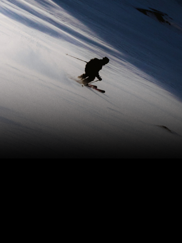 A skier in silhouette moves quickly down a snowy slope with visible ski tracks behind them.