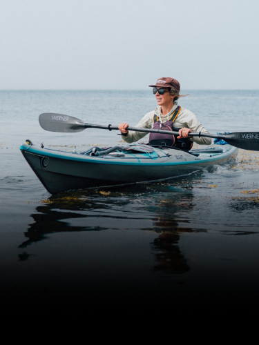 Person wearing a hat and sunglasses paddling a blue kayak on calm water near the shore, with the horizon visible in the background.