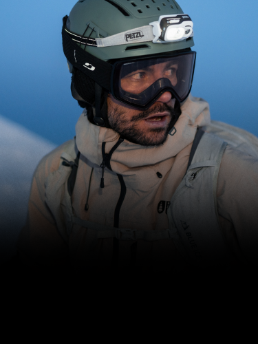 A person wearing a helmet with a headlamp, ski goggles, and a beige outdoor jacket, looking to the side against a blue background.