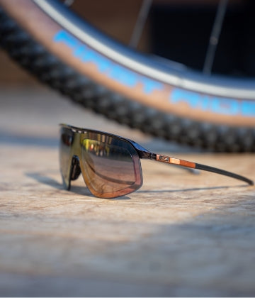 A pair of tinted sunglasses with thin frames rests on a wooden surface in front of a bicycle tire.
