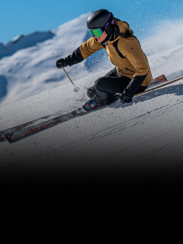 A skier in a brown jacket and helmet carves through snow on a sunny mountain slope.