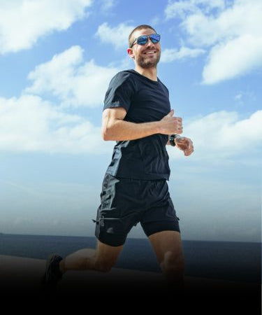 A man wearing sunglasses and black athletic gear is jogging outdoors under a blue sky with scattered clouds.