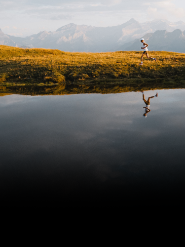 A person runs along a grassy hill near a calm body of water with mountains in the background and their reflection visible in the water.