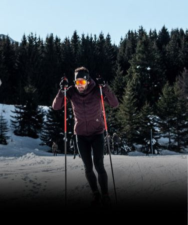 A person cross-country skiing on a snowy trail with a forest of evergreen trees in the background.