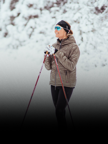 A person in winter clothing stands on snow with ski poles, surrounded by snow-covered trees.