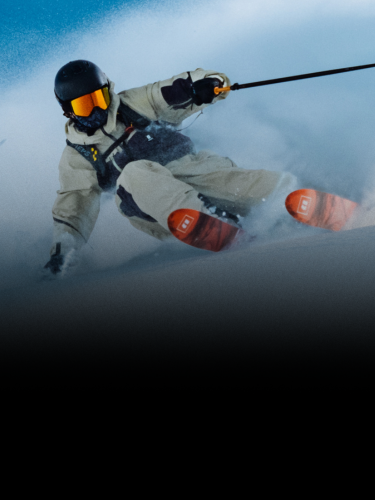 A skier in beige gear and orange boots makes a sharp turn on a snowy slope, kicking up powder, under a clear blue sky.