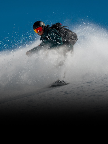 A person wearing a helmet and goggles snowboards downhill, kicking up powder against a clear blue sky.
