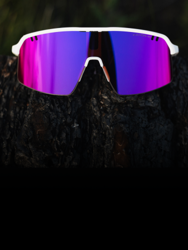 White sports sunglasses with mirrored pink-purple lenses resting on a dark, textured surface with blurred green foliage in the background.