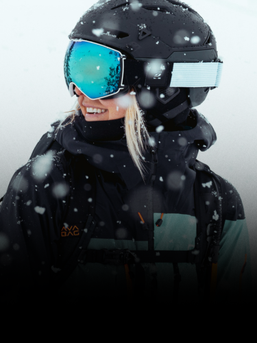 Person wearing a black ski helmet, blue reflective goggles, and ski jacket stands outdoors in falling snow, looking to the side and smiling.