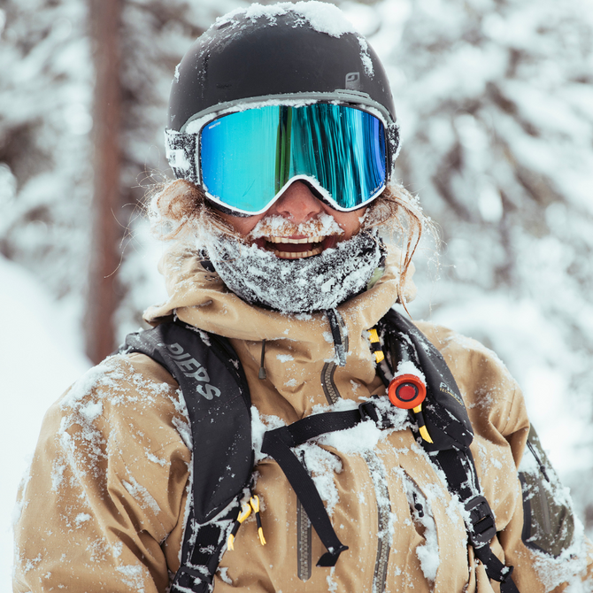 Person wearing a helmet, ski goggles, and winter gear stands outdoors in a snowy environment, with snow covering their clothing and face.