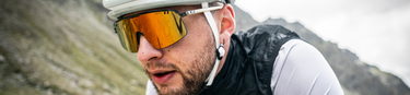 Close-up of a cyclist wearing a white helmet, large orange-tinted sunglasses, and a black vest, riding outdoors with blurred mountains in the background.