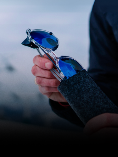 A person holds blue-lensed sunglasses, partially pulling them out of a gray felt case.