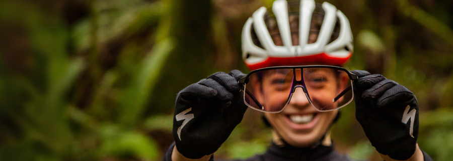 A cyclist wearing a helmet and gloves holds up a pair of sunglasses outdoors, smiling, with a blurred natural background.