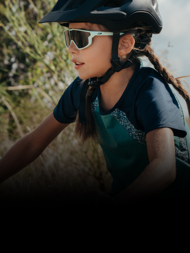 A young girl wearing a bike helmet, sunglasses, and a blue shirt rides a bicycle outdoors on a sunny day.