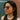 A woman in profile wearing round silver-framed sunglasses with reflective orange-brown lenses, photographed against a split green and brick wall background.